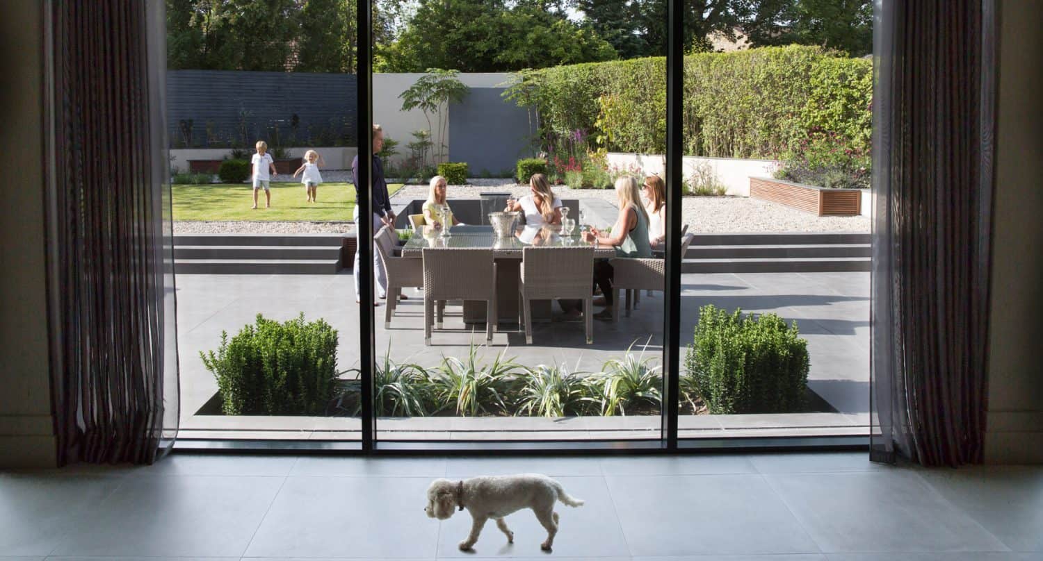 A small white dog walks inside by a glass door. Outside, several people sit at a patio table while children play on the lawn in a neatly landscaped garden on a sunny day.