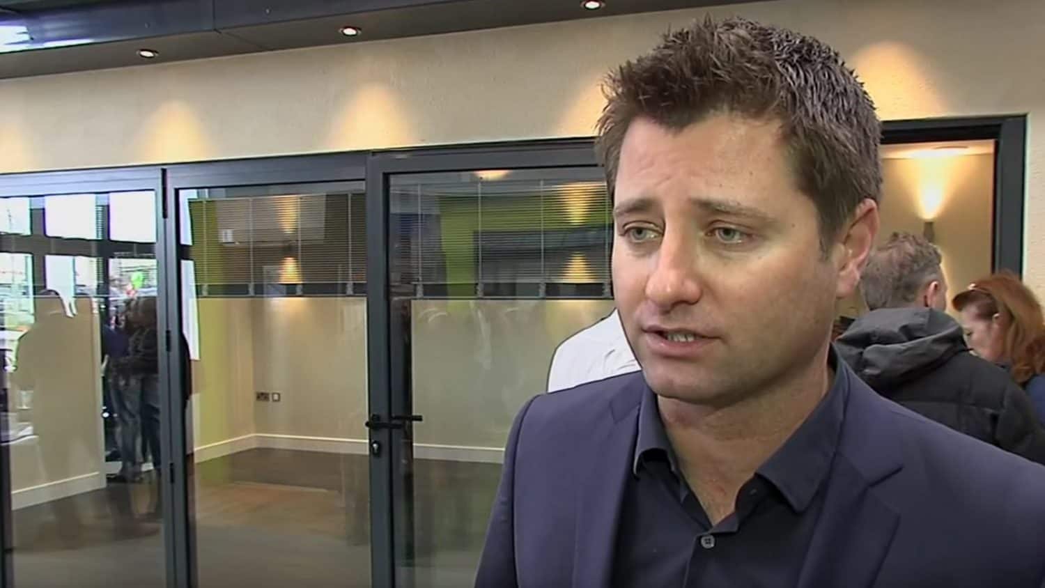 A man with short brown hair in a dark suit stands indoors, speaking, with glass bifold doors and several people blurred in the background.