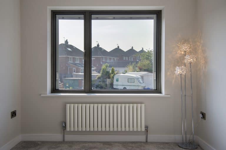 A bright room with a large window featuring bespoke glass solutions, offering views of houses, trees, and a white caravan. Inside, a radiator sits below the window and a modern floor lamp with decorative glass shades stands on the right.