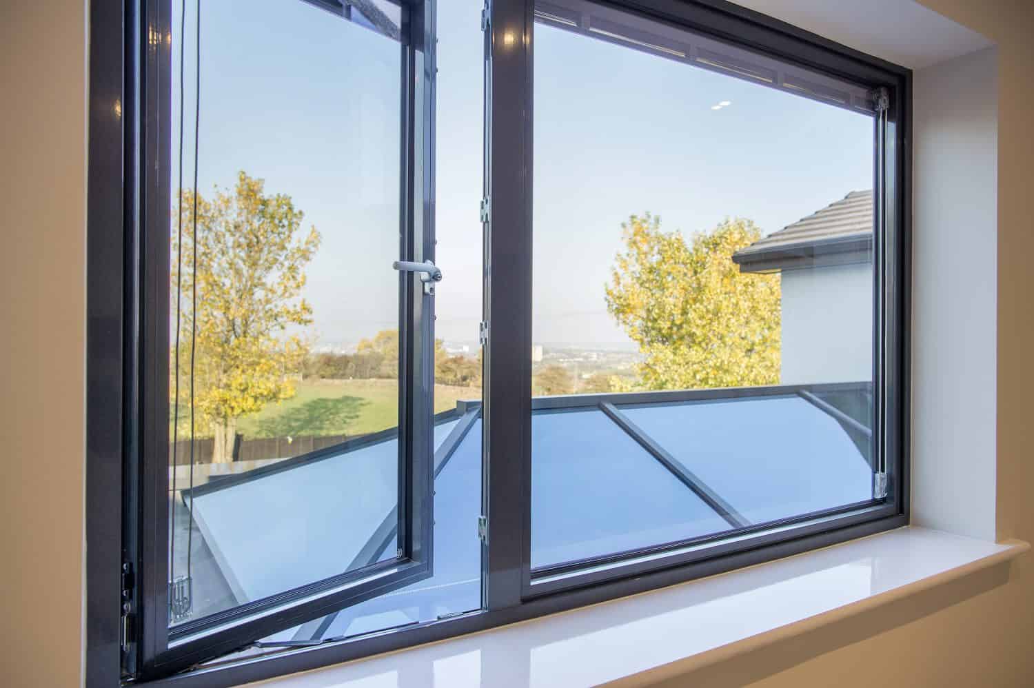 Open window with dark frame, showing a view of trees with autumn leaves, a distant landscape, and part of a roof featuring bespoke glass solutions. The bright daytime scene highlights elegant glass roofing outside.