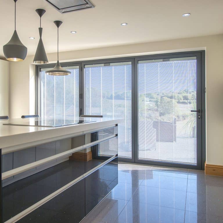 Modern kitchen with glossy black cabinets, a white countertop, and three pendant lights. Large sliding glass doors with sheer blinds let in natural light and feature bespoke glass solutions, offering a stunning view of greenery outside.