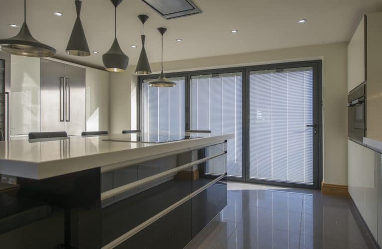 Modern kitchen with glossy black cabinets, a large white island, pendant lights, built-in appliances, and bifold doors letting in natural light. The space appears sleek and minimalistic with bespoke glass solutions enhancing its style.