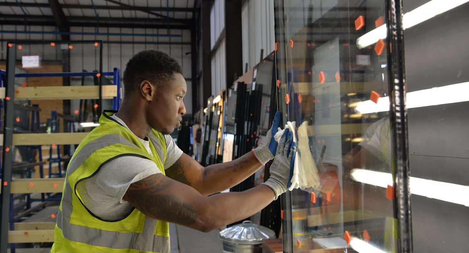 A worker in a high-visibility vest and gloves cleans a large glass pane in an industrial warehouse, surrounded by racks of glass destined for front doors and bespoke glass solutions, illuminated by bright overhead lights.