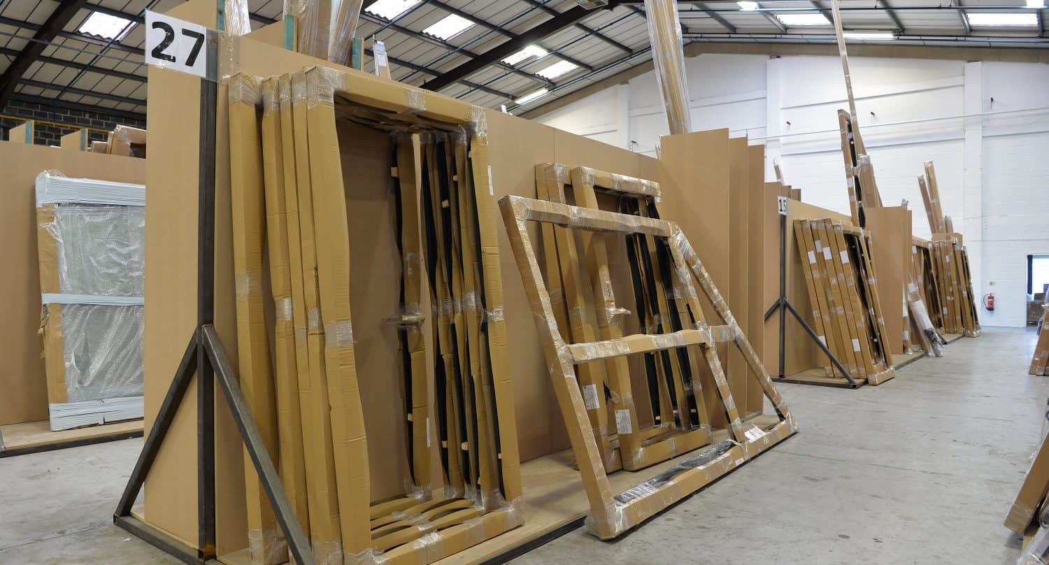 Large cardboard-wrapped frames and panels, including sliding doors and front doors, are stacked on metal racks in a spacious warehouse with a high ceiling. The racks are labeled with numbers, and more wrapped items are visible in the background.