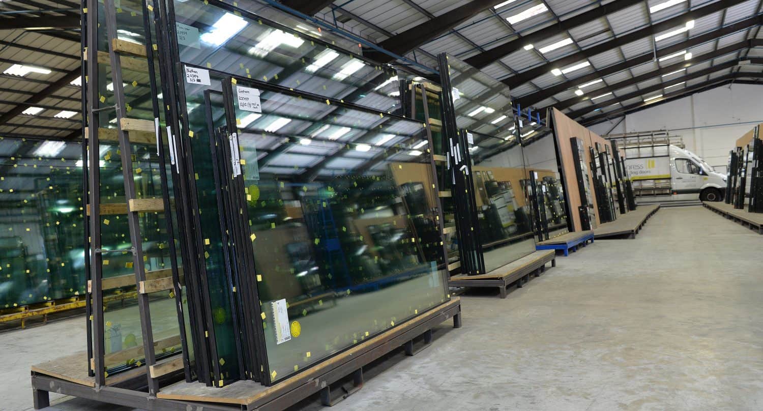 Large glass panels for sliding doors are stored vertically on wooden racks inside a spacious warehouse with a high ceiling. A delivery truck is parked in the background near the open entrance.