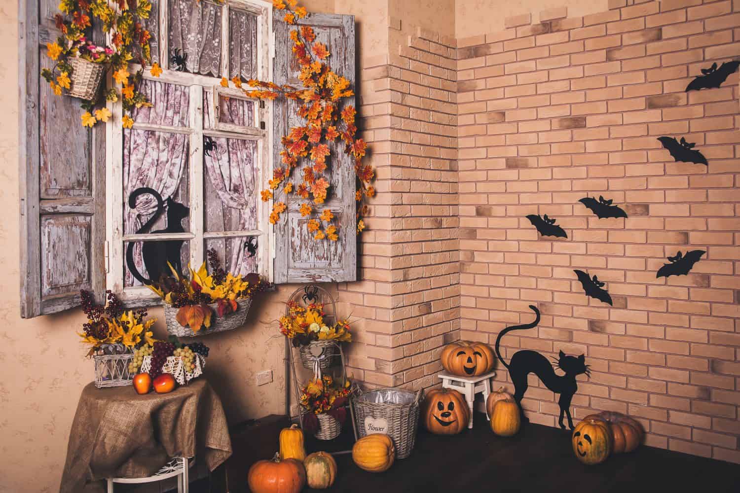 A cozy room decorated for Halloween with pumpkins, a black cat cutout, autumn leaves, and black bat silhouettes on a brick wall. Through stylish sliding doors, you can spot a window featuring a witch silhouette.