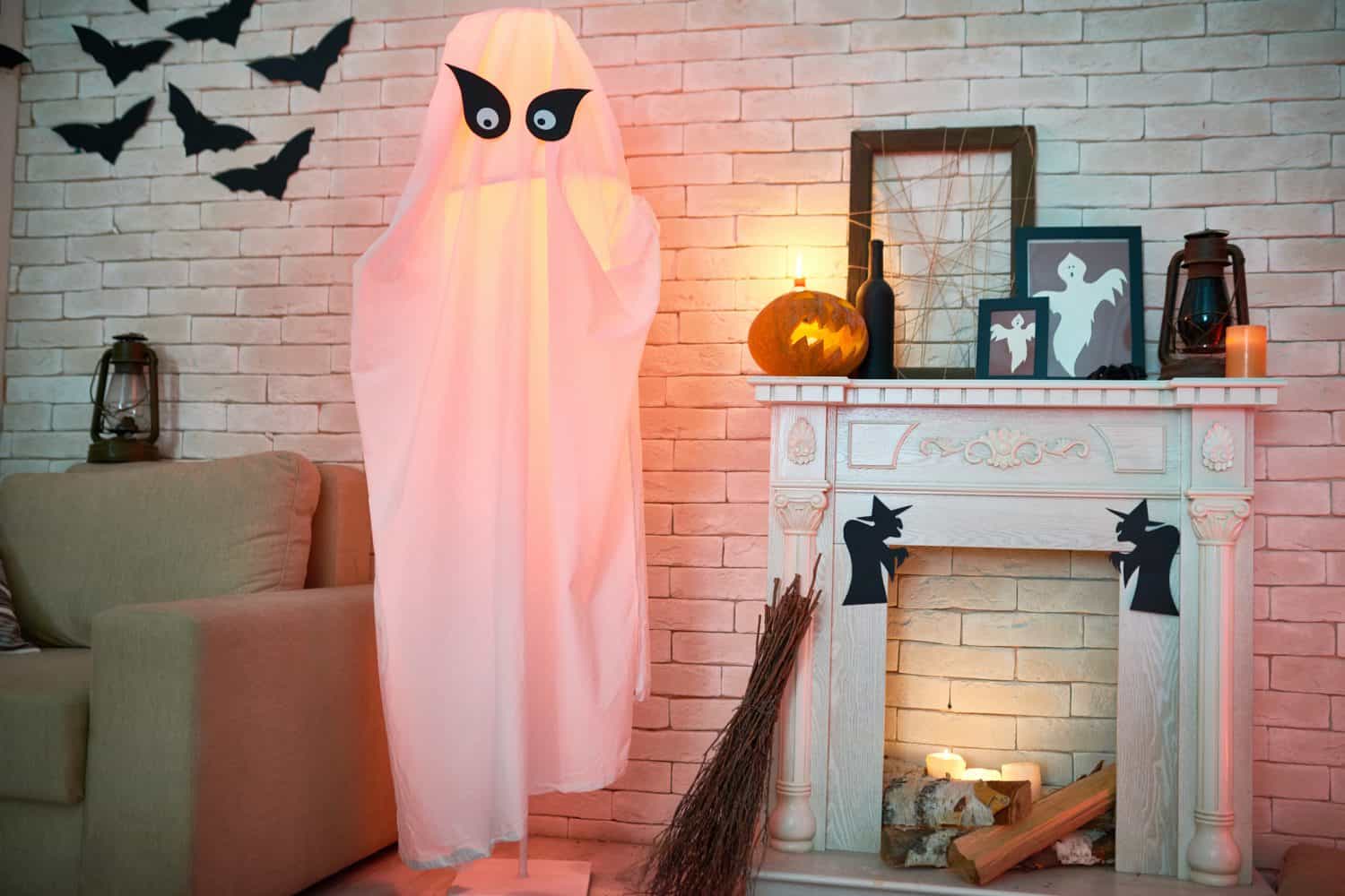 A Halloween-themed living room features a ghost decoration with big eyes, paper bats on the wall, a carved pumpkin, candles, and framed Halloween art by a white fireplace with sliding doors opening to the night.