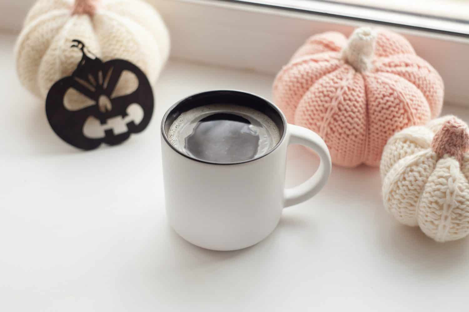 A white mug filled with coffee sits on a windowsill, surrounded by knitted cream and pink pumpkins, a small black paper jack-o-lantern, and sunlight streaming through bespoke glass solutions above.