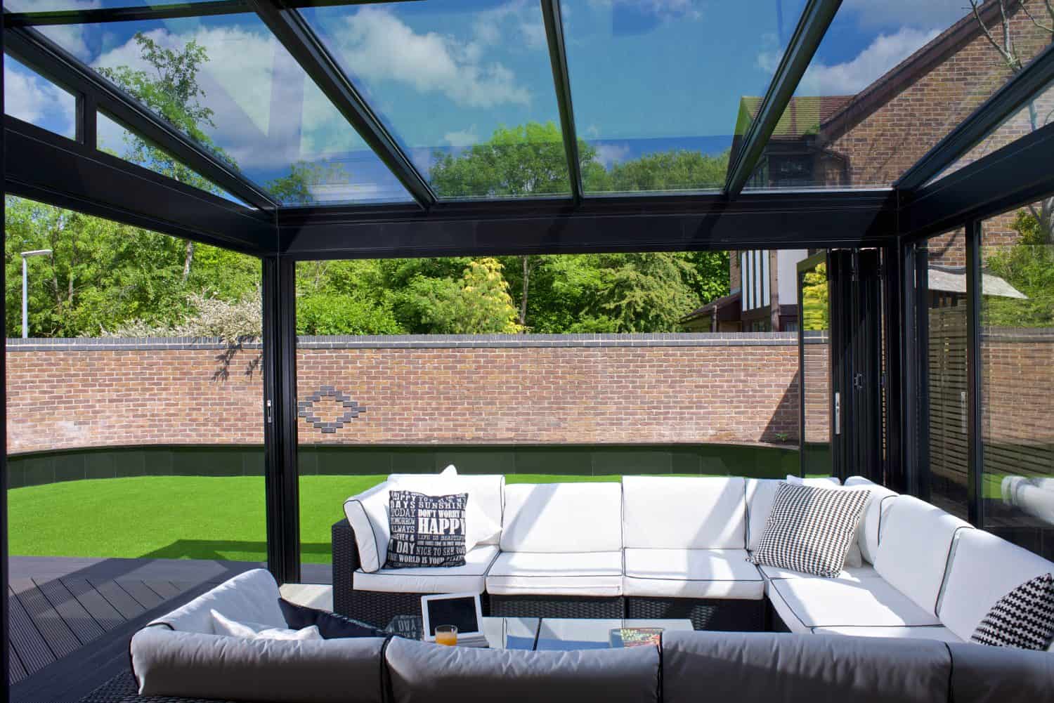 A modern glass conservatory with a white L-shaped sofa, throw pillows, and a coffee table sits beside a green lawn. Large sliding doors and a glass roofing reveal a brick wall and lush trees outside under a blue sky.