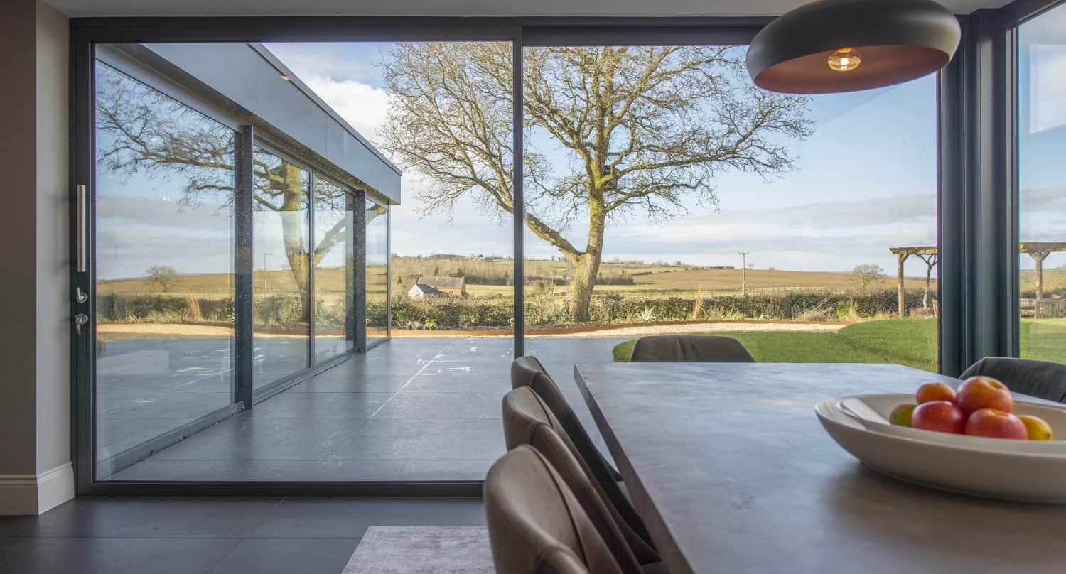 Modern dining area with a large sliding door—crafted using bespoke glass solutions—overlooking a patio, tree, and scenic countryside. A bowl of fruit sits on the table, with natural light filling the space beneath a pendant light.