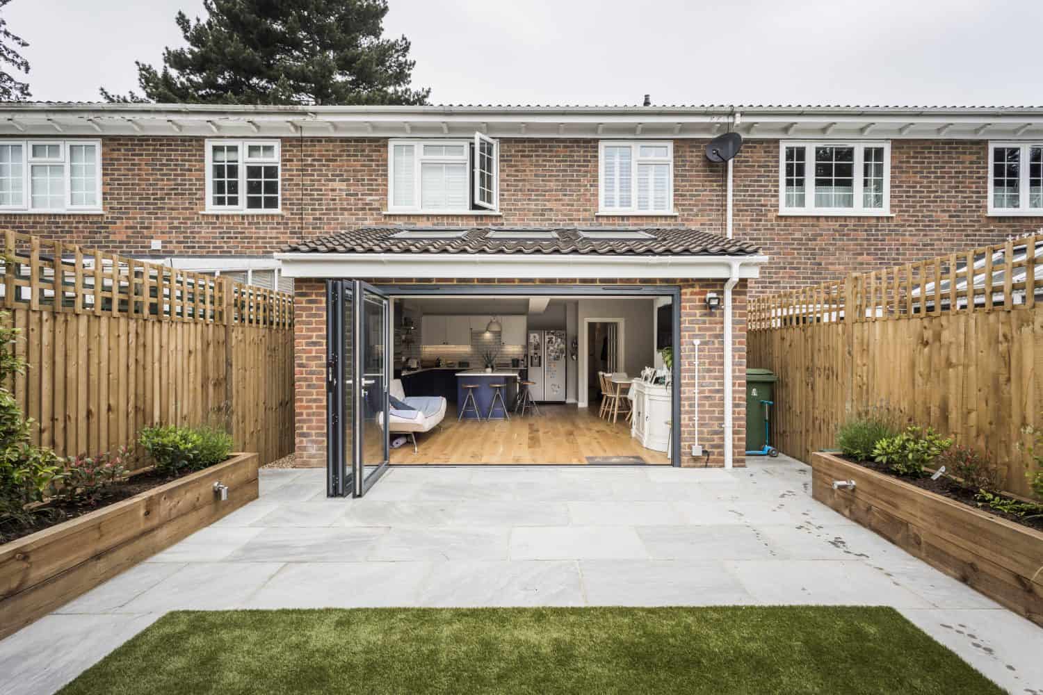A view of a modern brick house with bifold doors opening to a patio and small backyard, featuring fenced sides, raised garden beds, and a cozy, open-plan kitchen and living area visible inside.