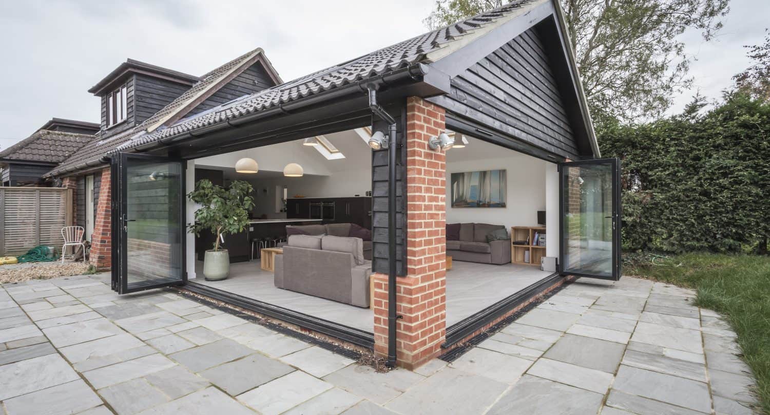 Modern home extension with large bespoke glass solutions, including bi-fold doors that open on two sides, revealing a spacious living area with sofas, pendant lights, indoor plants, and artwork. The patio is paved with stone tiles.
