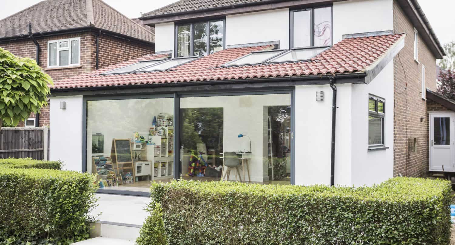 A modern house with a red-tiled roof and bespoke glass solutions, featuring large glass sliding doors opening onto a patio. Inside, childrens toys and books are visible. The garden has neatly trimmed hedges and a potted plant.