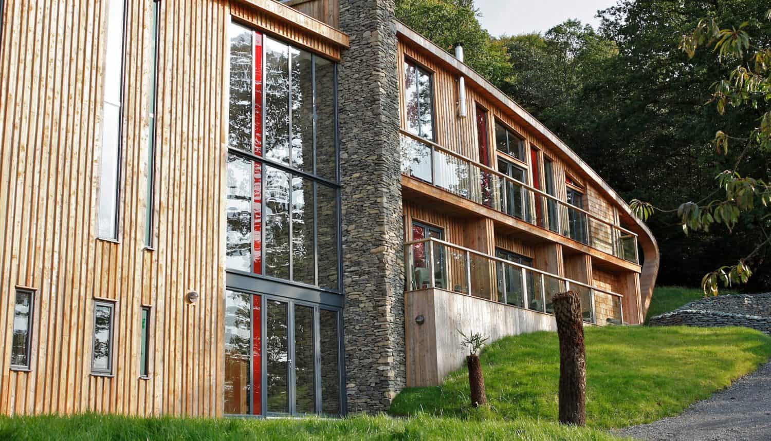 Wooden and stone-clad modern building with bespoke glass solutions, large sliding doors, and a curved design, set on a grassy slope next to trees and a gravel path.
