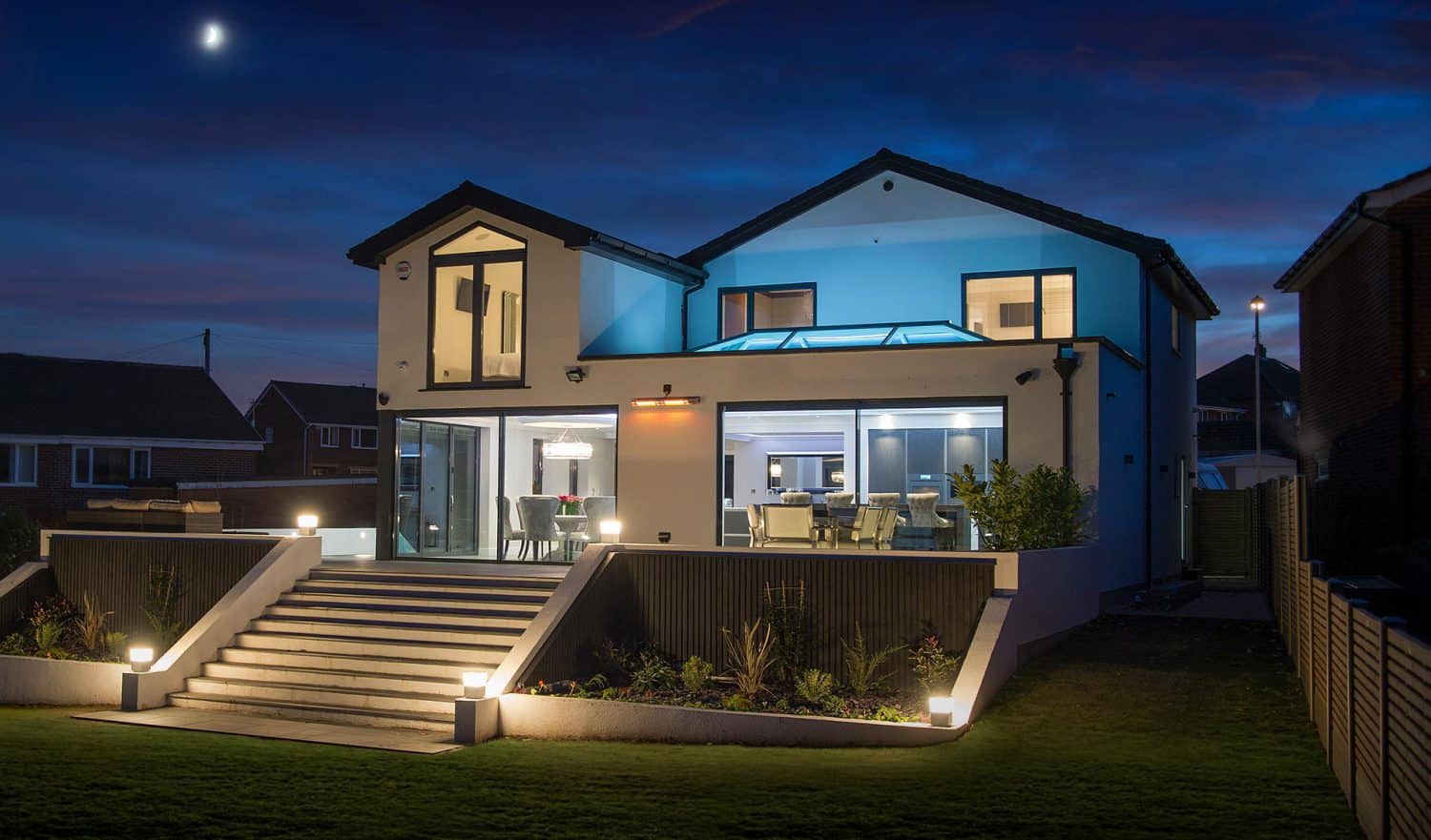 Modern two-story house with large windows and outdoor lighting, illuminated at night. Wide steps lead to stylish front doors, while interior lights reveal a contemporary dining and living area. A crescent moon graces the sky above.