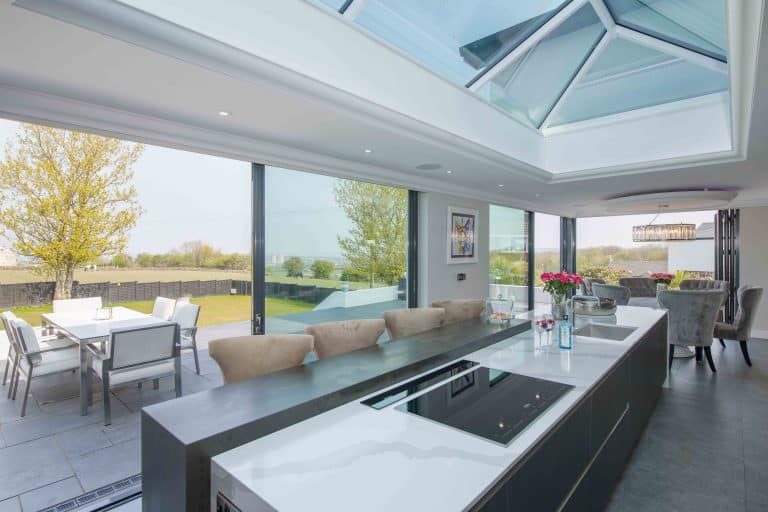 Modern kitchen with a large island, built-in stovetop, and sink. Bifold doors open to a patio with outdoor dining, letting in natural light. Skylights and trees are visible outside. Roses sit on the counter.
