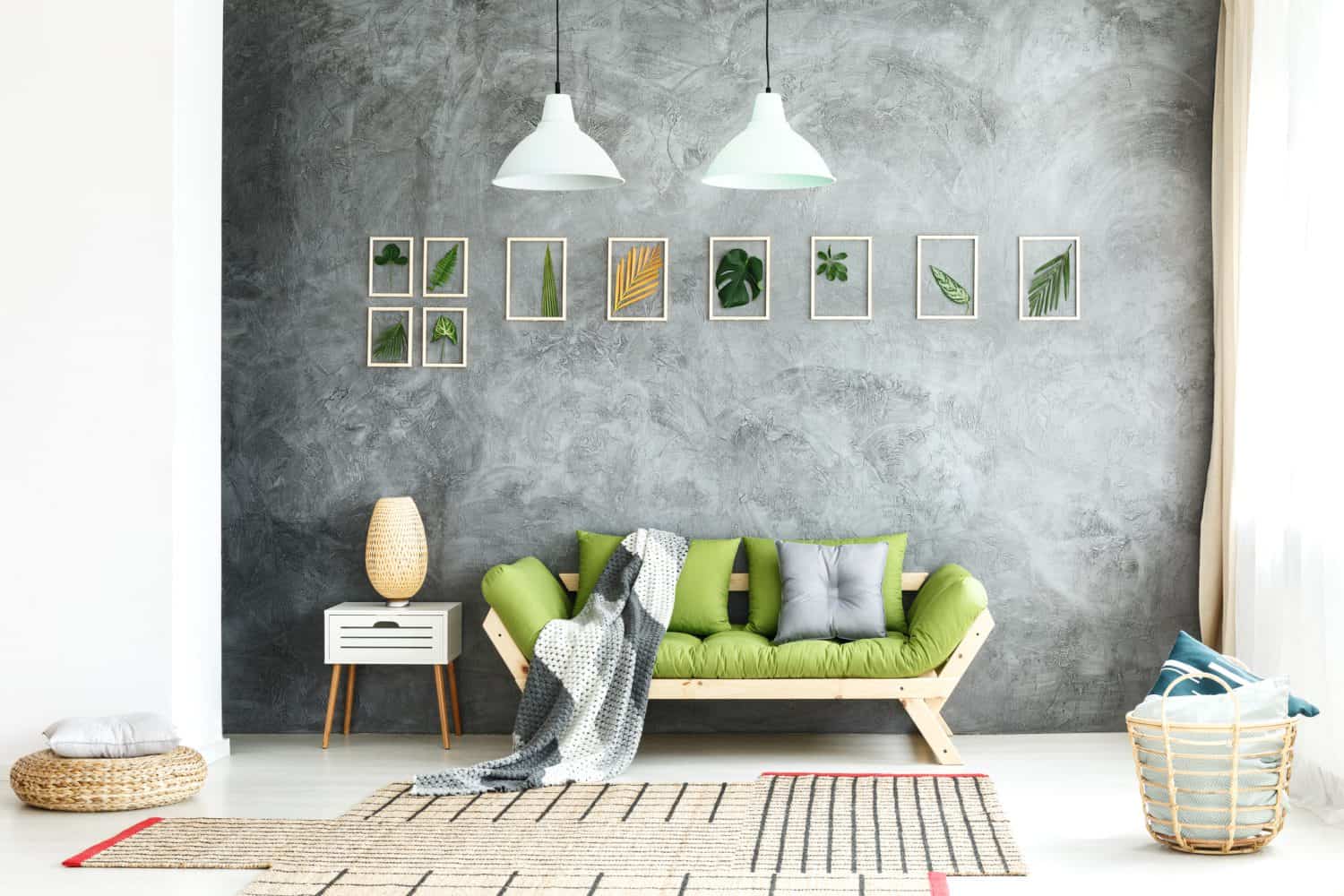 Knitted blanket on wooden sofa, comfy pouf and basket with soft pillows in cozy living room interior with natural green accents