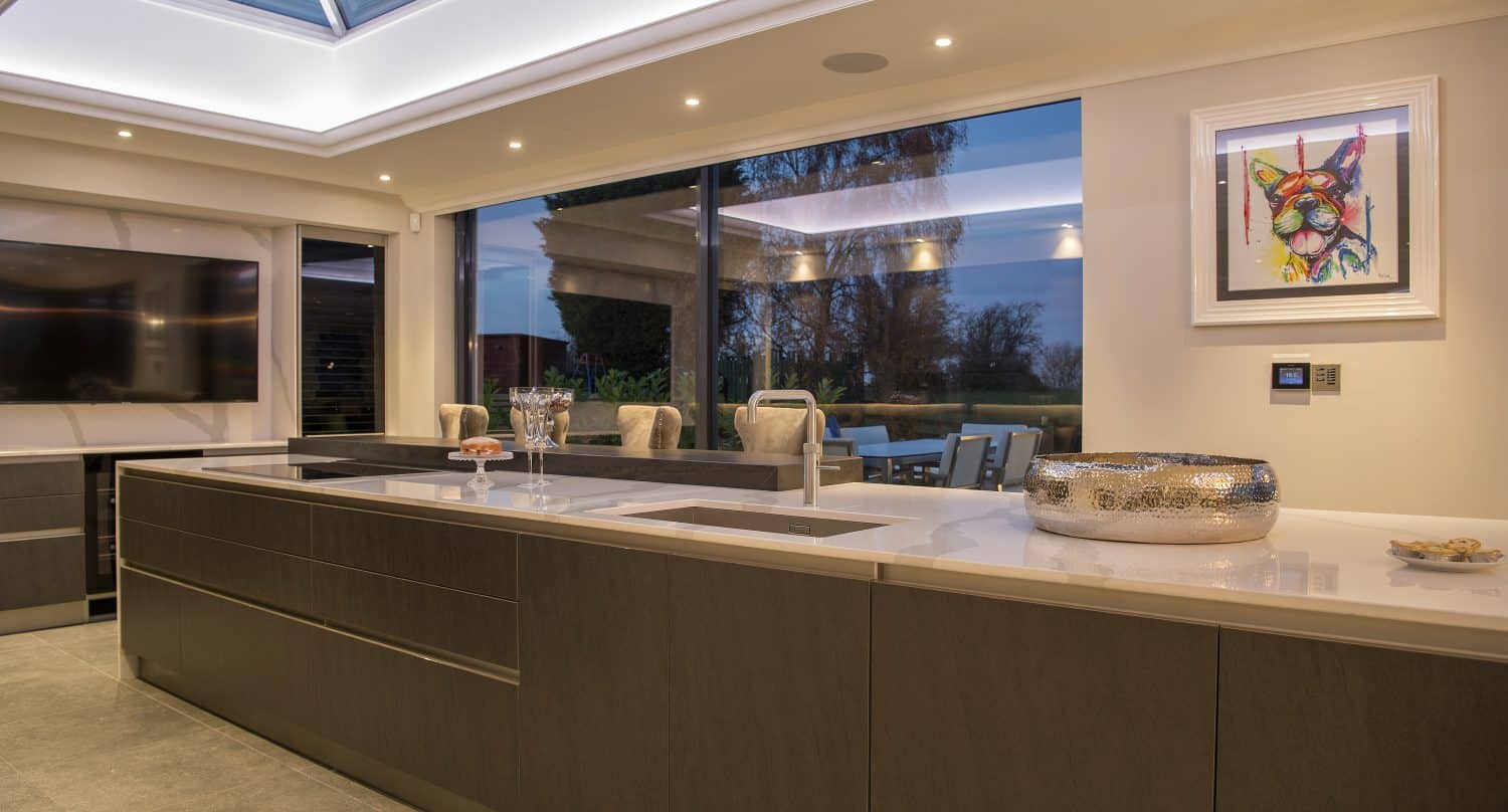 Modern kitchen with large island, sleek dark cabinets, white countertops, and a skylight ceiling created with bespoke glass solutions. Bifold doors open to an outdoor dining area, while artwork and a silver bowl decorate the space. Evening light is visible outside.