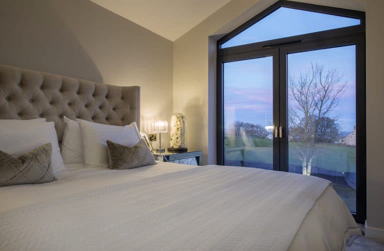 A cozy bedroom with a large bed, tufted headboard, and two decorative pillows sits beside a lamp-lit table. Bespoke glass solutions in the large doors reveal a tree and colorful evening sky just outside.