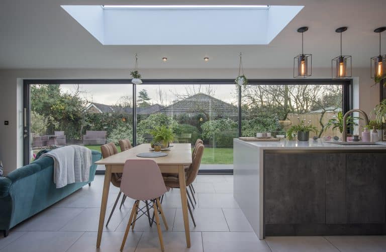 Modern kitchen-diner with a skylight, wooden table and pastel chairs, green sofa with a white blanket, indoor plants, and bespoke glass solutions such as large sliding doors opening to a lush garden.