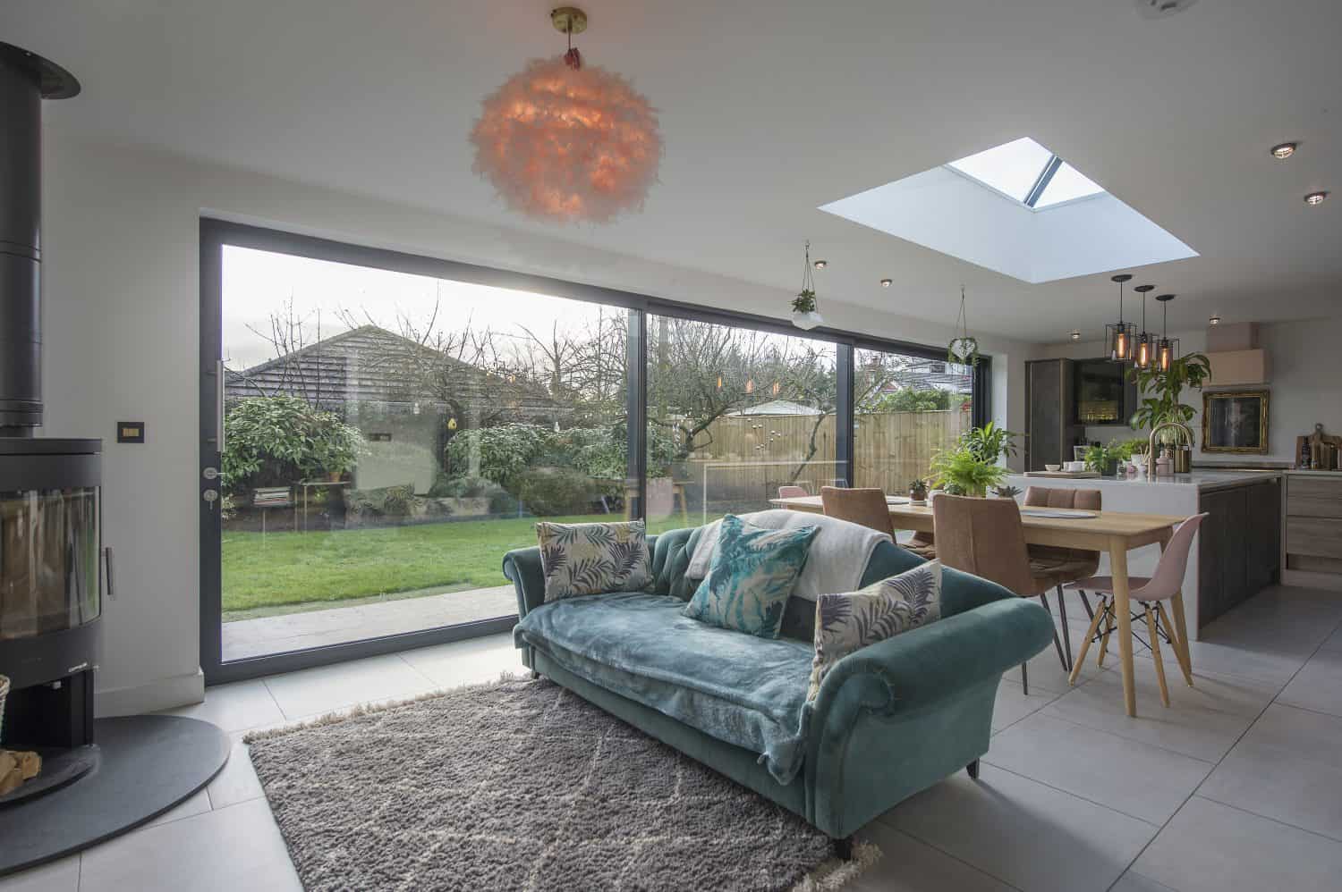 A modern open-plan living and dining area features a teal velvet sofa, a fluffy rug, bespoke glass solutions like bifold doors overlooking the garden, a skylight, dining table with pink chairs, and lush indoor plants.