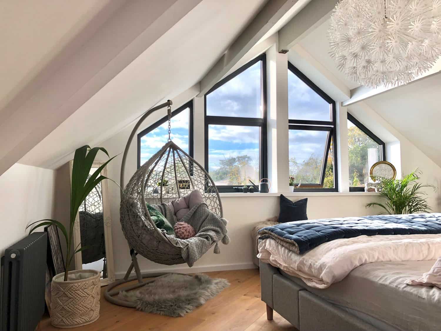 A cozy attic bedroom with a large triangular window, glass roofing above the grey bed, plants, and a hanging chair with cushions and a blanket, all under white sloped ceilings with exposed beams.