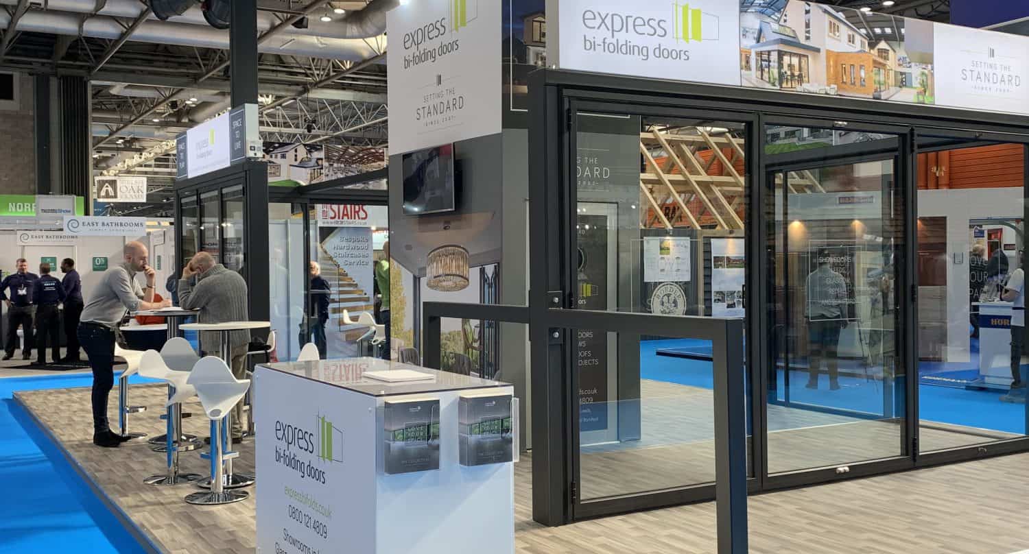 An exhibition booth for Express Bi-Folding Doors features large sliding door displays, a counter with brochures, white stools, and signage under bright lighting on a blue carpeted floor at a trade show.