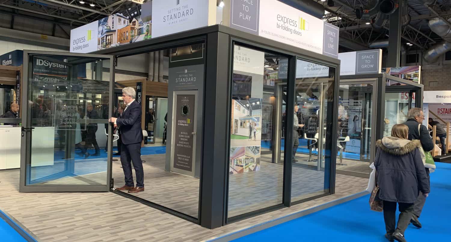 A trade show booth displays glass doors, sliding doors, and windows. A man in a suit stands inside, opening a glass door. People walk by on a blue carpet in a large indoor exhibition hall with high ceilings.