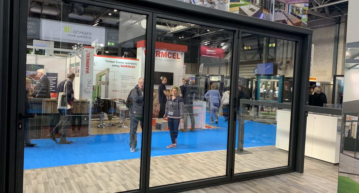 People are walking inside a trade show or exhibition hall with stands and banners. The photo is taken through large sliding doors, revealing a blue carpet and various displays visible in the background.