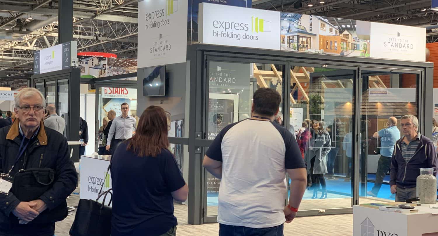 People walk around an indoor trade show featuring a large glass display from Express Bi-Folding Doors showcasing bespoke glass solutions, with bright lighting overhead and informational booths in the background.