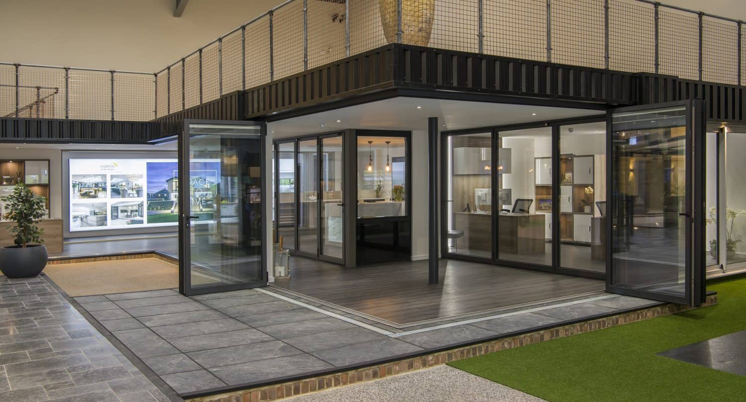 Modern indoor showroom featuring a corner glass room with folding doors and bespoke glass solutions, surrounded by artificial grass, tiled floors, and display walls with windows and doors. A balcony with metal railing is visible above.