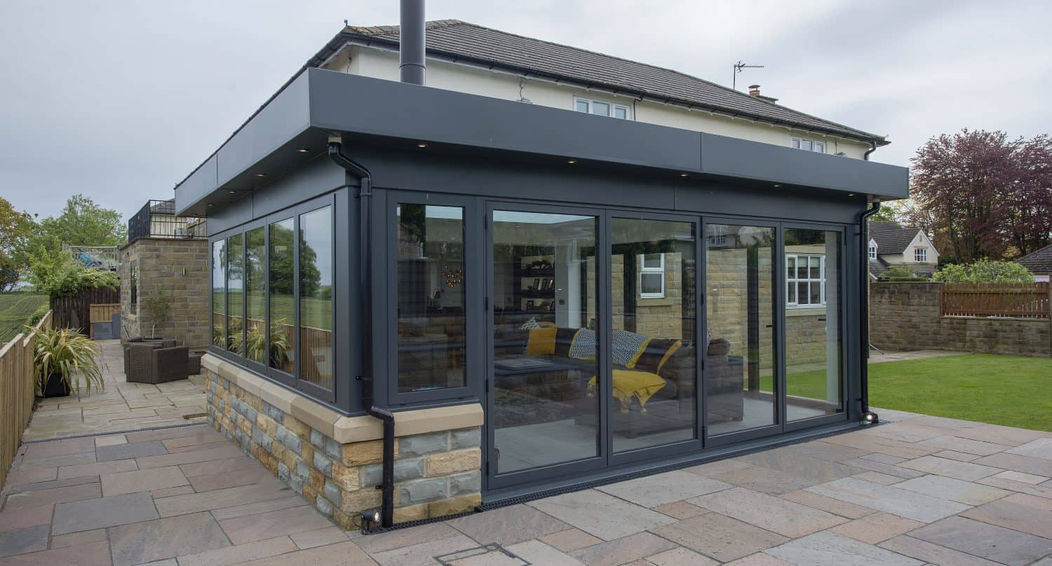 A modern glass and steel sunroom extension with bespoke glass solutions and sliding doors, attached to a stone house, overlooks a large paved patio and a grassy yard bordered by a stone wall and trees.