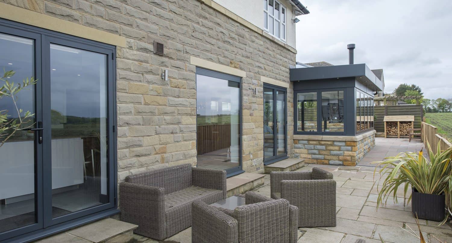 A modern stone patio with wicker chairs and a glass table sits outside a house featuring large sliding doors and windows. Potted plants add charm, while a covered BBQ area enhances the inviting atmosphere.