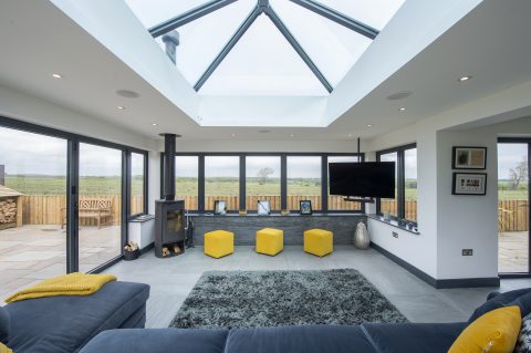 Modern living room with large glass windows, a skylight ceiling, and countryside views. The space features yellow stools, a gray rug, a sofa, TV, framed photos on a shelf, and sliding doors leading to the visible outdoor patio.