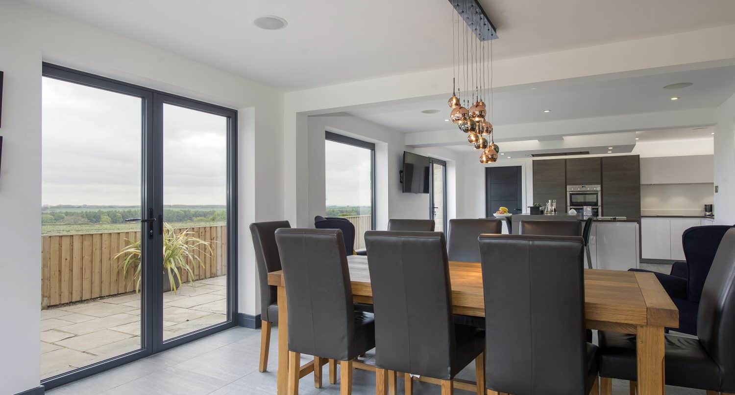 Modern dining area with a wooden table, eight dark leather chairs, and pendant lights. Large bespoke glass sliding doors and windows provide views of an outdoor patio and greenery. A kitchen is visible in the background.