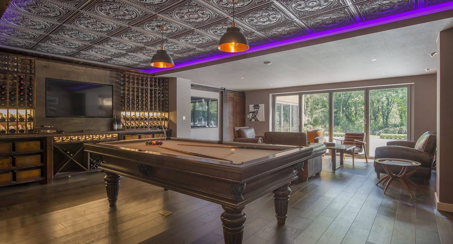 A stylish game room with a dark wood pool table, ornate patterned ceiling, pendant lights, wine racks, TV, seating area, and large windows with sliding doors overlooking a green outdoor space.