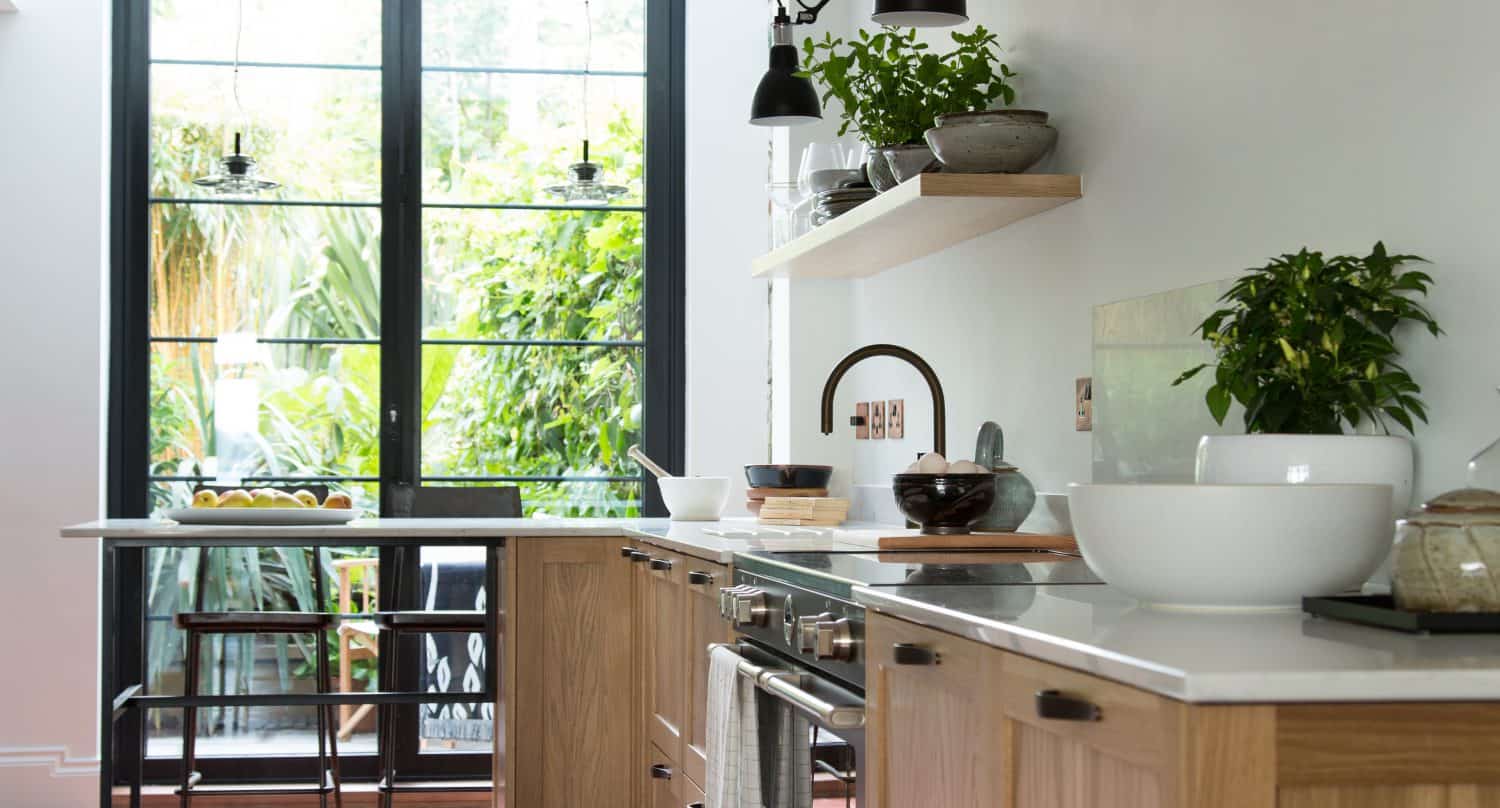 Modern kitchen with wooden cabinets, white countertops, potted plants, and black wall-mounted lights. Large windows and sliding doors at the end let in natural light and offer a view of green foliage outside.