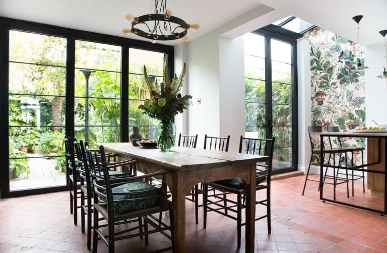 Bright dining area with large windows, wooden table and black chairs, bifold doors opening to green plants outside, floral wallpaper on one wall, and a vase of flowers as the centerpiece on the table.