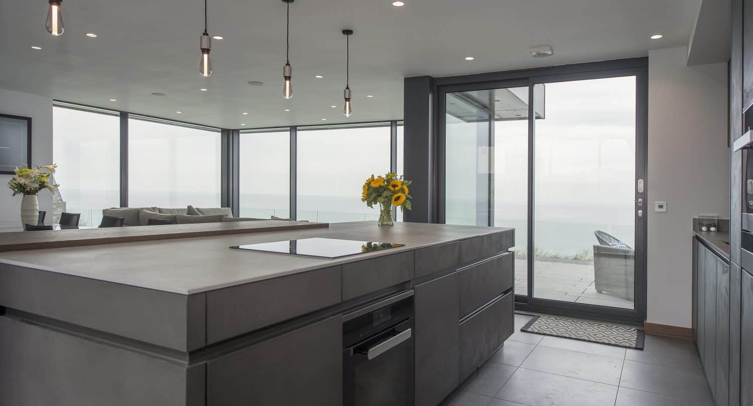 Modern kitchen with dark cabinets and island, pendant lights above, large windows offering ocean views, and bespoke glass solutions like a bifold door leading to a patio. Sunflowers in a vase decorate the countertop.