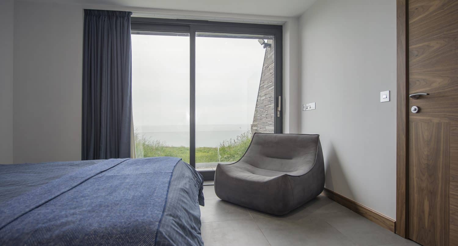 A modern, minimal bedroom with a blue bedspread, gray lounge chair, wooden sliding doors, and a large window with dark curtains, offering a view of grass and the sea outside on a cloudy day.