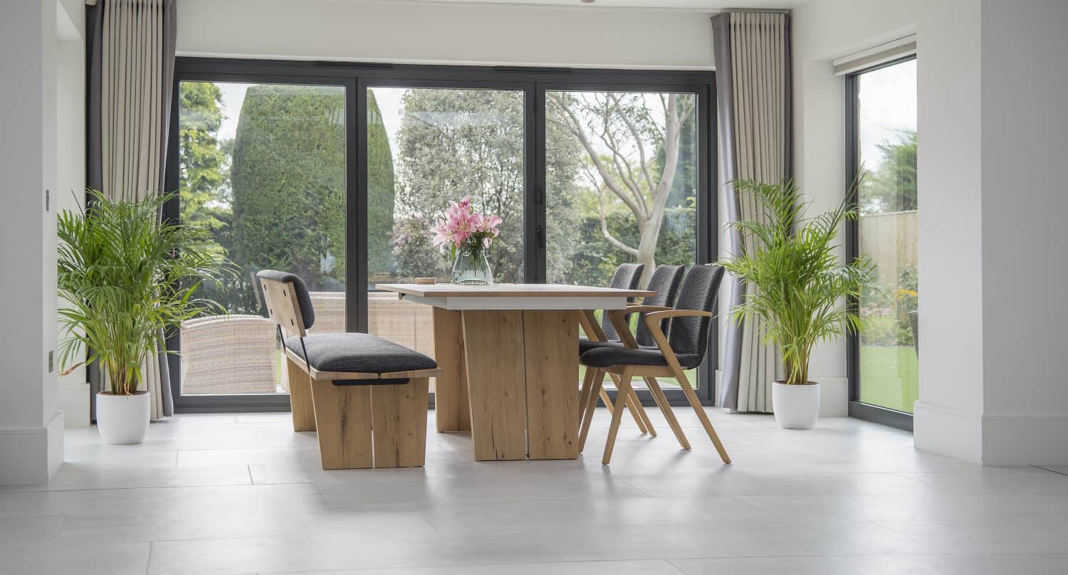 Modern dining room with a wooden table, bench, and chairs, flanked by potted plants. Large sliding doors and windows let in natural light and offer views of a garden with trees and greenery outside.