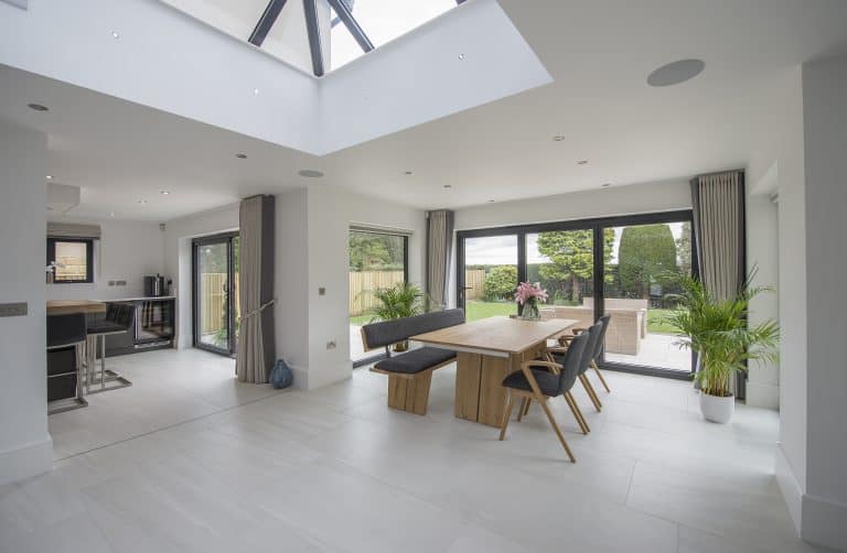 A modern, bright dining area with large windows and bifold doors, a wooden table with six chairs, plants, and views of a sunny patio and garden. Open layout connects to the kitchen as natural light fills the space.