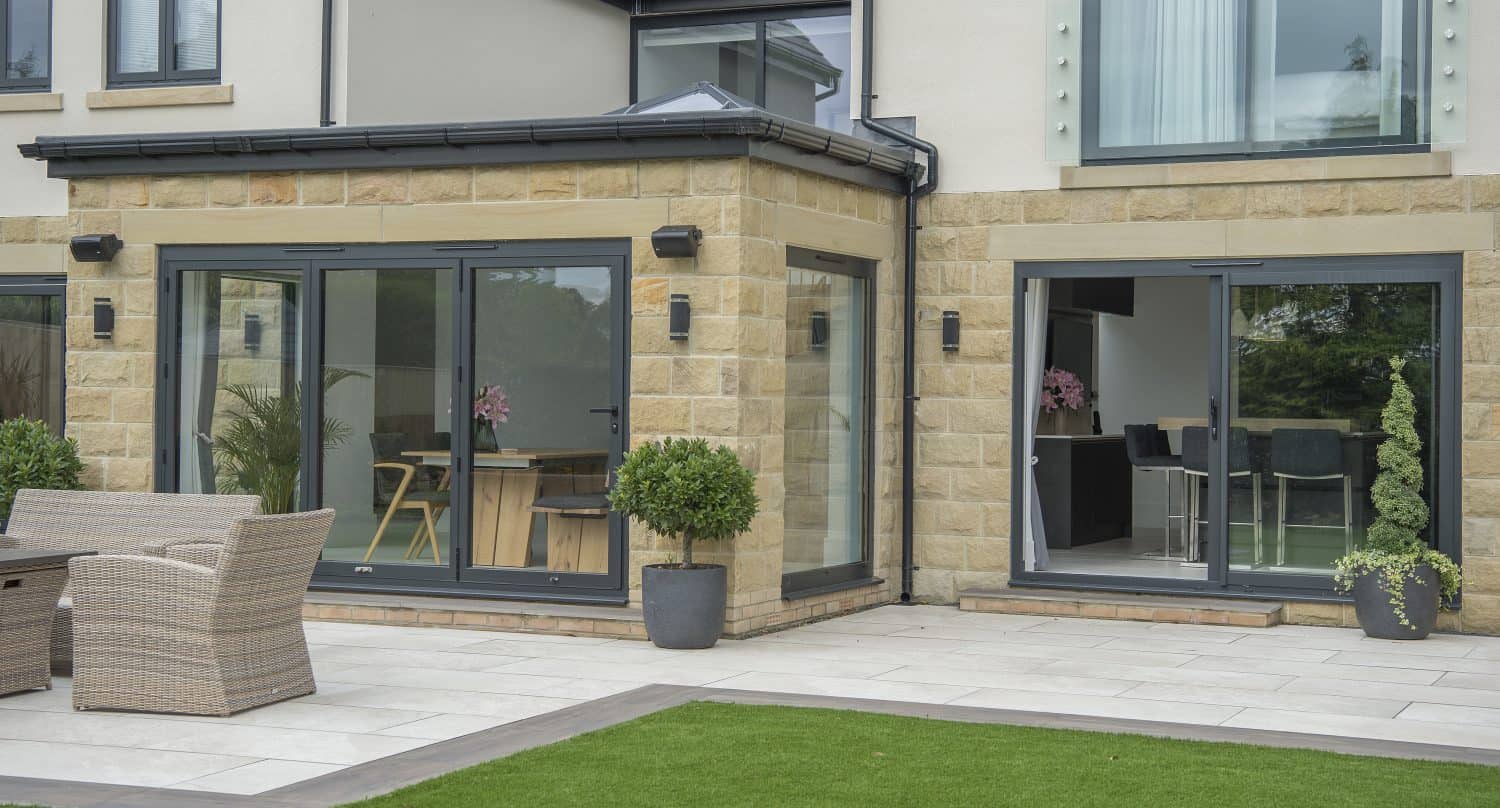 Modern house with large glass bifold doors, stone and cream exterior, and a patio area with wicker furniture. Potted plants line the patio, while a lush green lawn stretches out in the foreground.