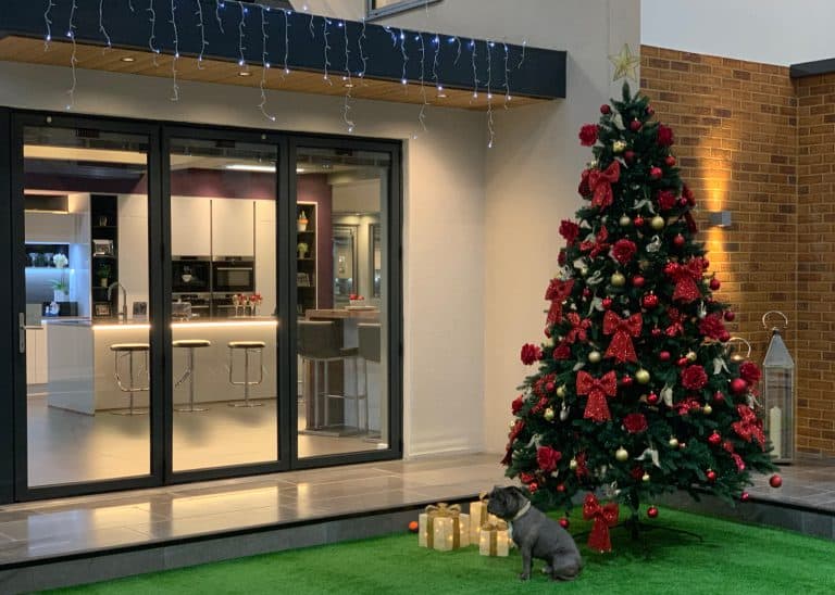 A decorated Christmas tree with red ornaments and bows stands on green artificial grass near wrapped gifts and a small dog, outside a modern house with large sliding doors and string lights.
