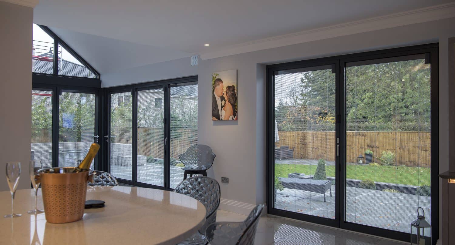 Modern dining area with sliding doors opening to a patio and garden. A table with a champagne bottle and glasses is in the foreground, and a framed photo hangs on the light gray wall. Outdoor seating is visible through the doors.