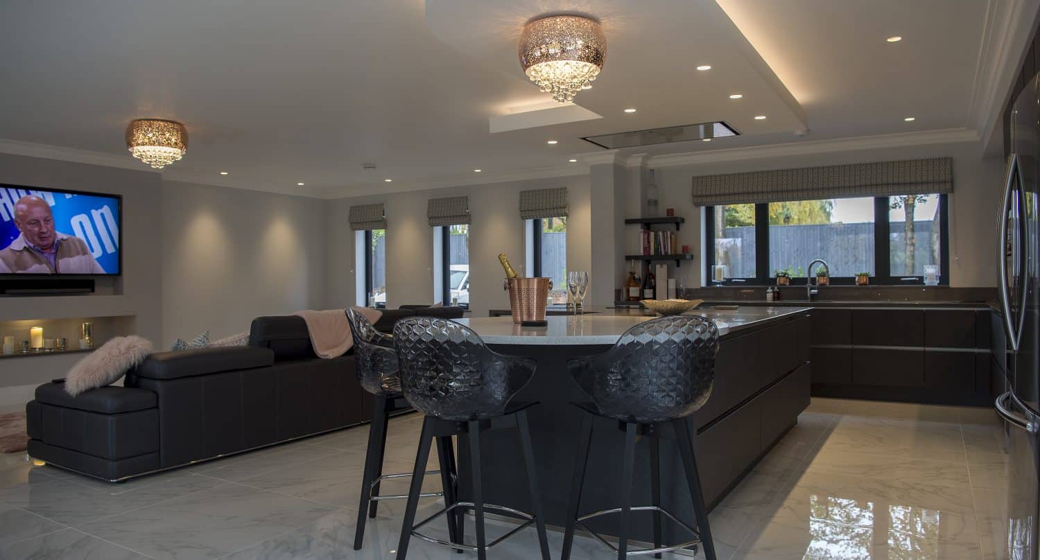 Modern open-plan kitchen and living area with black island, stylish bar stools, marble flooring, elegant light fixtures, large sofa, wall-mounted TV, and bifold doors letting in natural light.