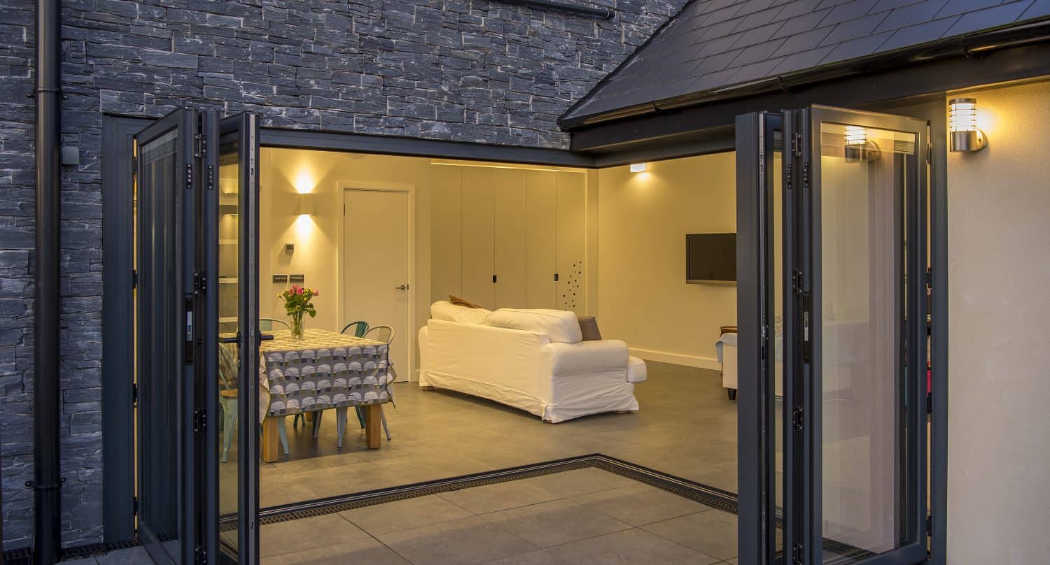 Modern home with large bifold doors open to a patio, revealing a well-lit living space with a white sofa, dining table with a patterned cloth, and stone wall exterior.