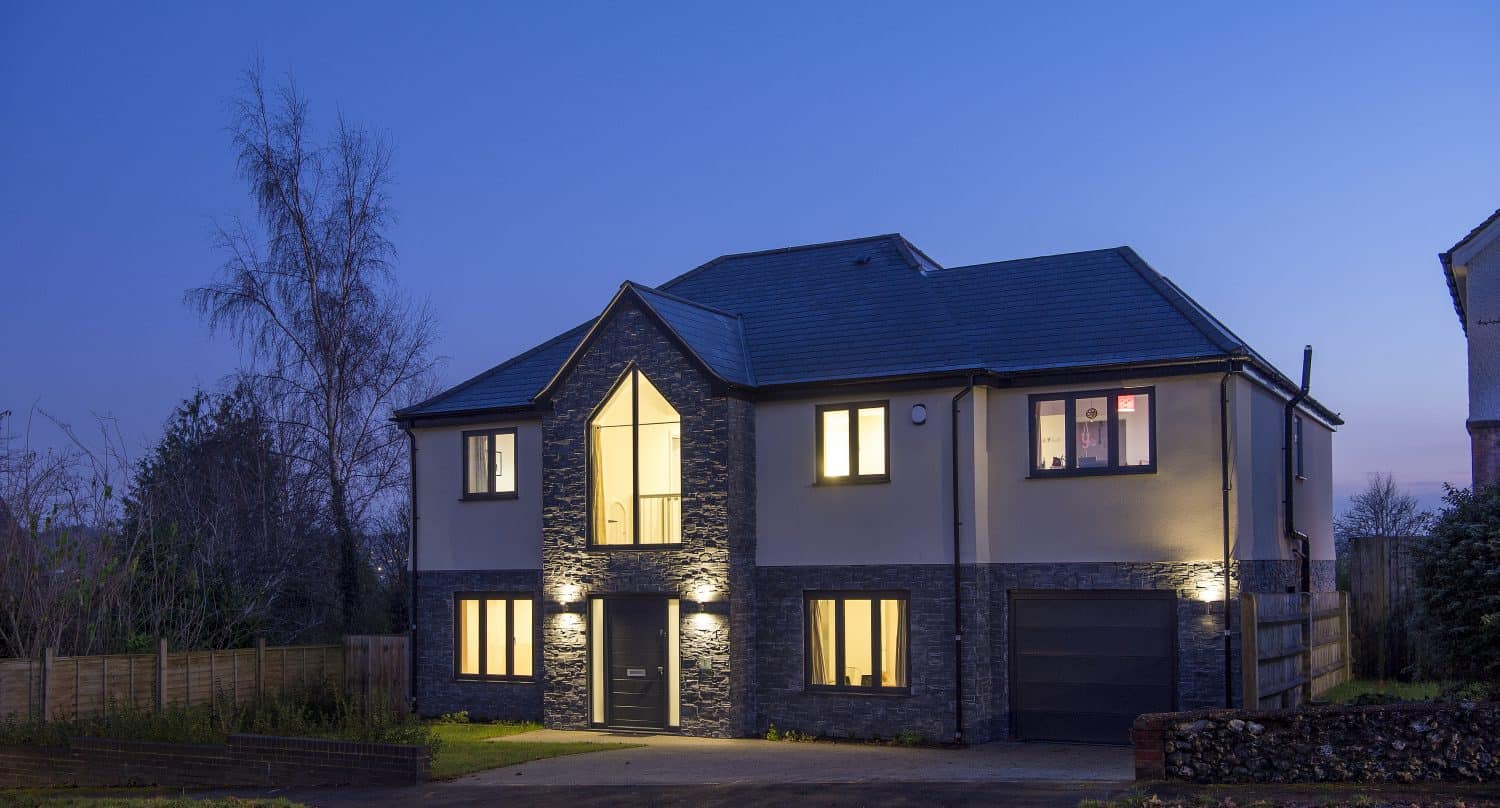 A modern two-story house with illuminated windows and stone accents features bespoke glass solutions in its front doors, photographed at dusk under a clear sky with a crescent moon. A driveway and small front lawn are visible.