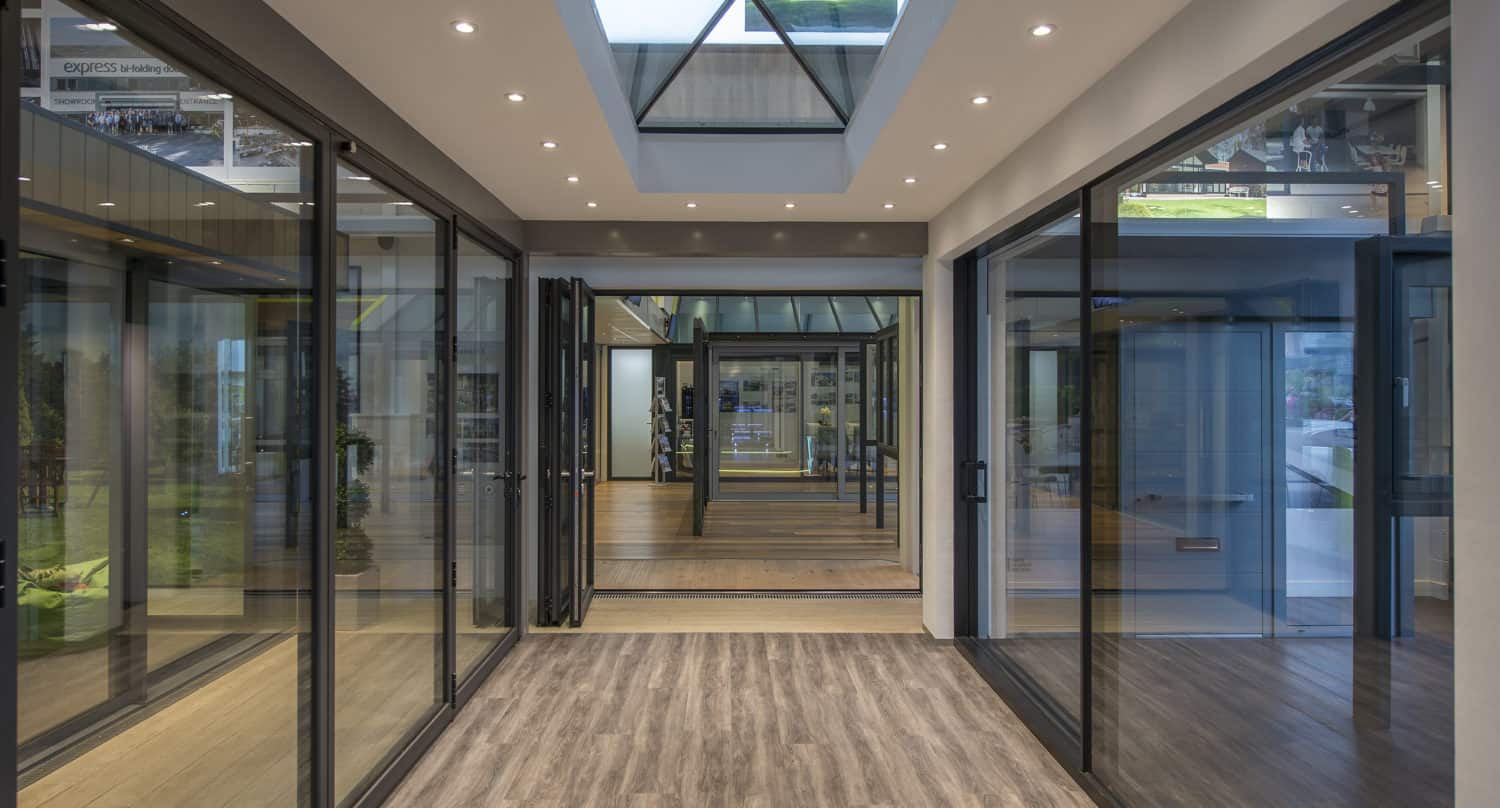 Modern showroom hallway featuring large sliding doors and wood-style flooring, illuminated by recessed ceiling lights and a skylight. Multiple reflections from the front doors create a spacious, open atmosphere.