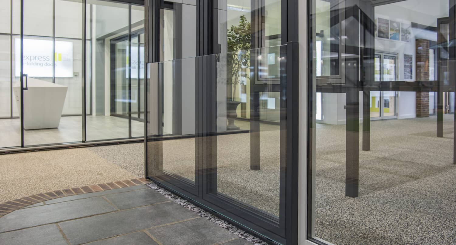 Modern office interior with bespoke glass solutions, featuring large glass walls and doors dividing rooms. The flooring transitions from dark tiles to light carpet, and a plant is visible in the background.